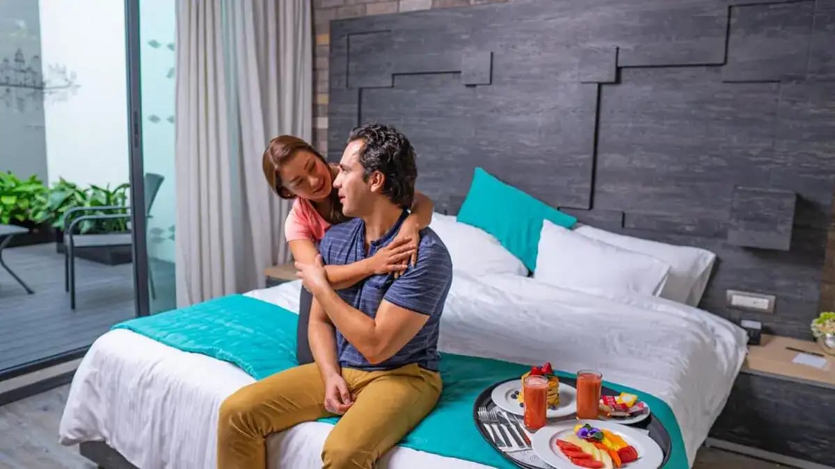 Pareja en la habitación Master Suite del Hotel Punto MX con una vista a la Ciudad de México. Tienen el desayuno en la cama.